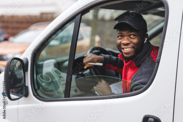 Fototapeta Physical working - package delivery. Positive smiling African-American man in black baseball hat sitting on driver's seat of a white van. View through vehicle window. High quality photo