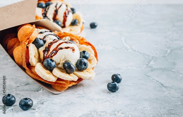 Fototapeta Delicious bubble waffle with ice cream, banana and blueberry on gray marble background.