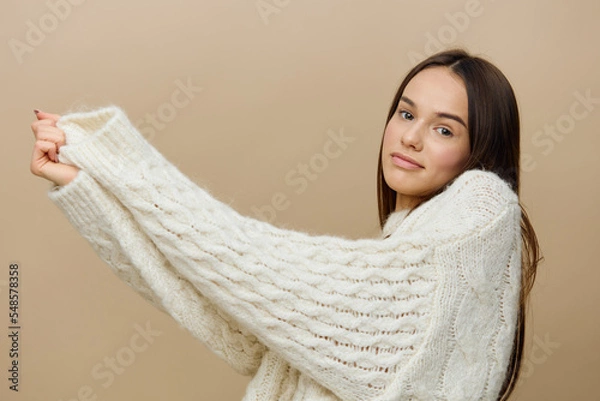Fototapeta a cheerful woman standing sideways on a light brown background in a white sweater raised her hands and pulled them aside, stretching the sleeve
