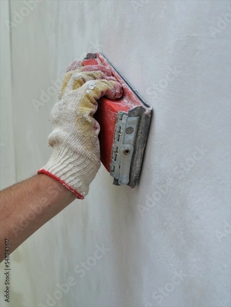 Fototapeta hand sander in a red case in the builder's glove hand close-up, in the process of finishing the plaster on the wall before painting or wallpapering, smoothing the roughness of the plastered wall