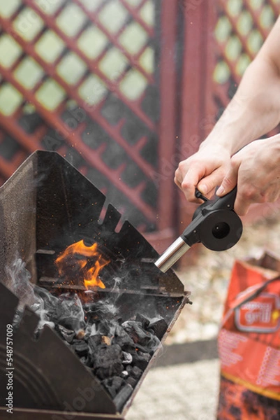 Fototapeta Man's hand holding and using air blower to fire up charcoal in a metal tray, portable grill barbecue. BBQ set up.