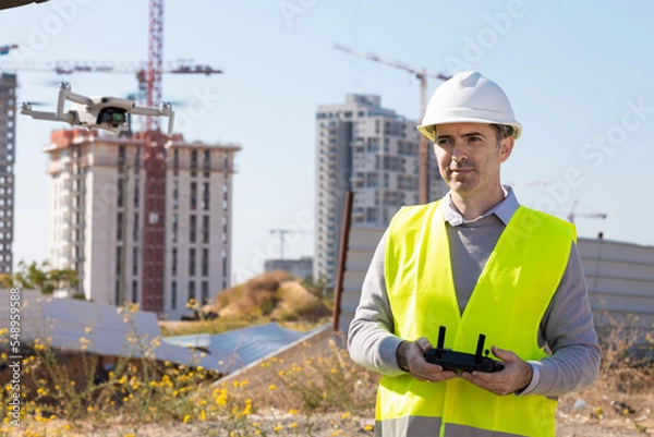 Fototapeta Man launches a quadcopter. An engineer flies a drone next to a construction site. Concept - construction observation with a drone.  Construction cranes on sunset sky