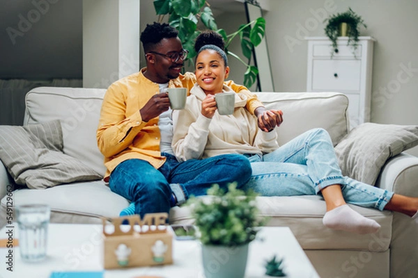 Fototapeta Multiracial couple relaxing on a couch at home and drinking coffee