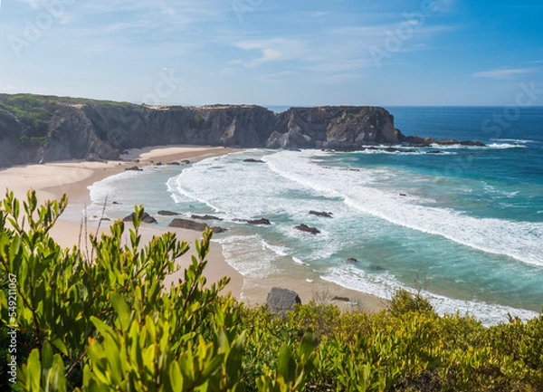 Fototapeta View of empty Praia dos Machados beach with ocean waves, cliffs and stones, wet golden sand and blurred green vegetation at foreground. Rota Vicentina coast, Odemira, Portugal.