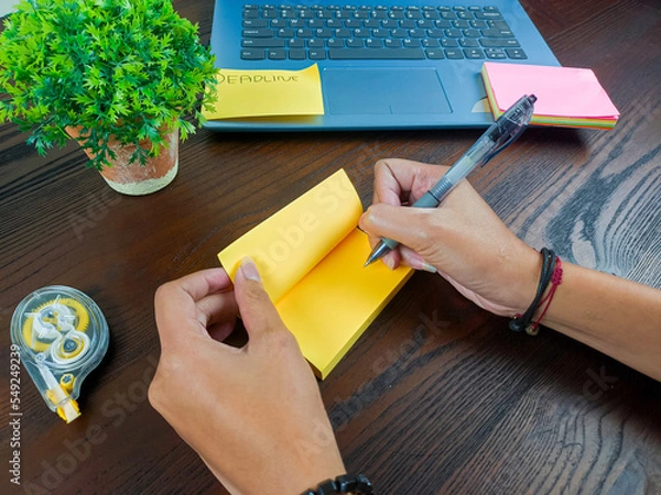 Fototapeta women write in yellow notes, mock up yellow notes. Women's hands write notes on yellow note paper in the background of the office desk workspace from the top view.