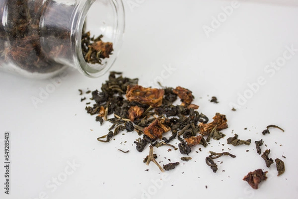 Obraz green tea leaves with pieces of fruit poured out of a glass bottle on a white background