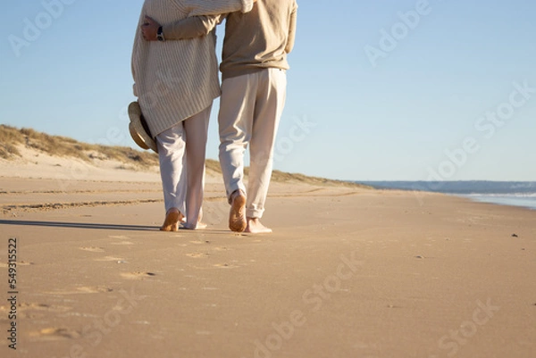 Fototapeta Romantic couple enjoying vacation at seaside while walking along beach and embracing. Back view of middle-aged man and woman spending holiday time together. Coastline background. Love, leisure concept