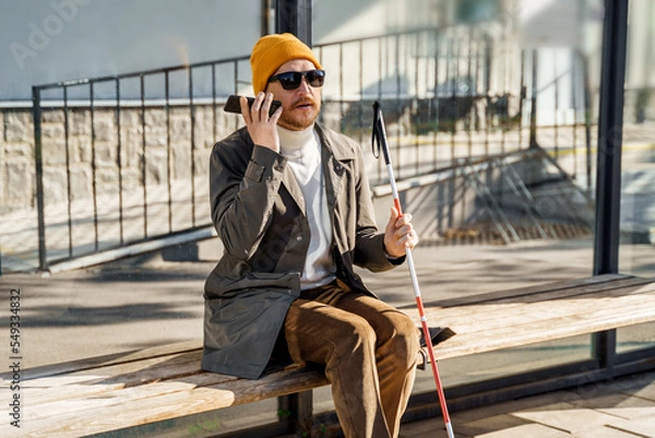 Fototapeta Blind person with a walking stick sits on a bench at a public bus stop uses a smartphone