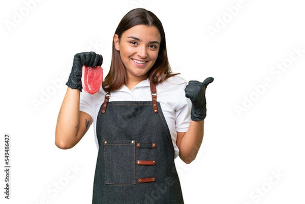 Fototapeta Butcher woman wearing an apron and serving fresh cut meat isolated on green chroma background pointing to the side to present a product