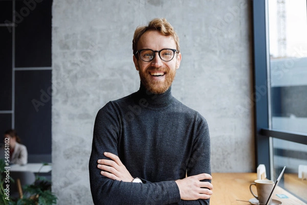 Obraz A young red-haired guy, a programmer or an entrepreneur in glasses, in a stylish cafe Looks at the camera and smiles. Freelancer works remotely. Online communication. small business.