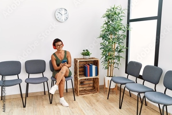 Fototapeta Young hispanic woman listening to music sitting on chair at waiting room