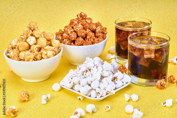 Obraz three bowls of spicy, caramelized and salted popcorn accompanied by 2 glasses of soda with ice on a yellow background with copy space for advertising.