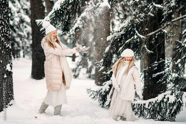 Fototapeta Handsome mother and daughter are having fun outdoor in winter time. Playing with snow in forest between snowy trees
