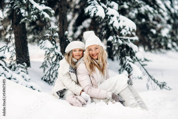 Fototapeta Handsome mother and daughter are having fun outdoor in winter time. Sitting on snow in forest between snowy trees