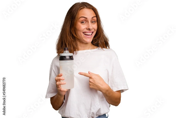 Fototapeta Young caucasian sport woman holding a bottle of water isolated smiling and pointing aside, showing something at blank space.
