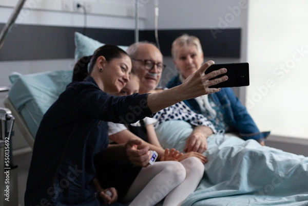 Fototapeta Accompanying hospitalized elderly man for routine checkups capturing cell phone snapshots at geriatric medical tower. Young woman taking selfie with sick father in clinic rest room.