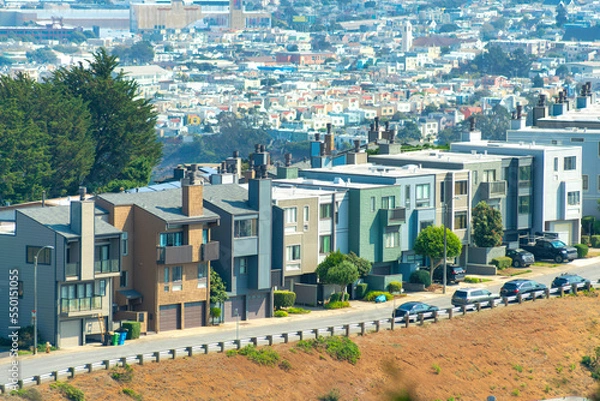 Obraz Aerial shot of multicolored row of houses with road in front and dry grassy hill and downtown city in sun background