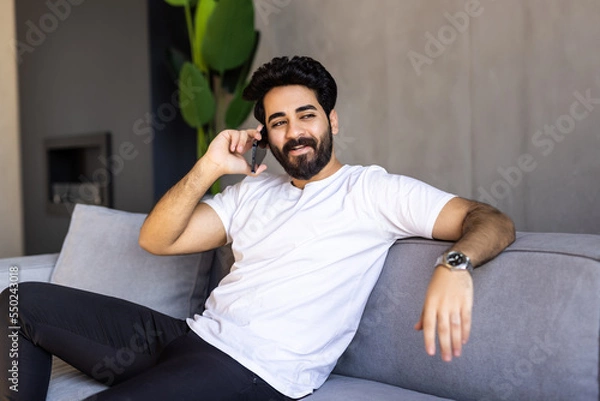 Fototapeta Happy emotional arab guy having phone conversation at home, got good news, sitting on sofa in living room.