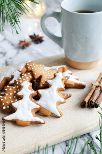 Fototapeta Festive cookies decorated with white icing on a wooden cutting board with a hot drink in a blue cup on a marble background with lights. Homemade ginger and cinnamon Christmas cookies. 
