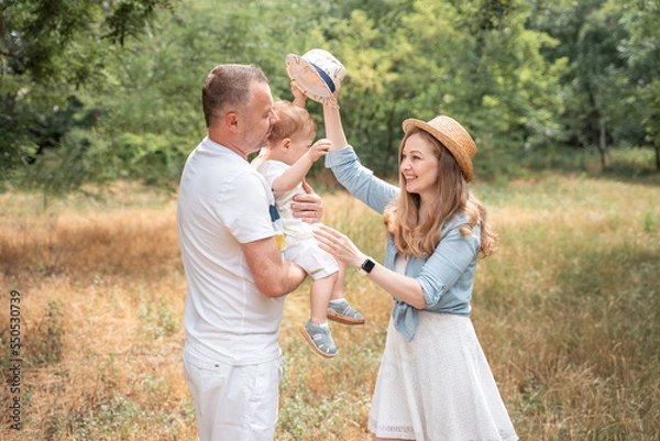 Fototapeta Parents are playing with their son in the clearing, mom is trying to put on a hat for the baby