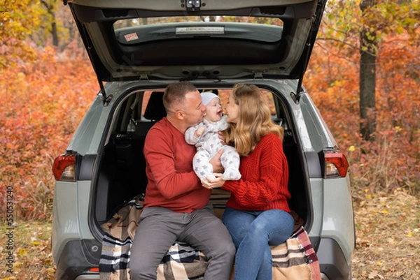 Fototapeta Parents in red sweaters sit in the trunk of a car and hug their baby