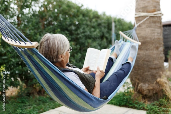 Fototapeta Smiling senior good-looking grey hair woman wearing glasses while reading in hammock in the summer garden