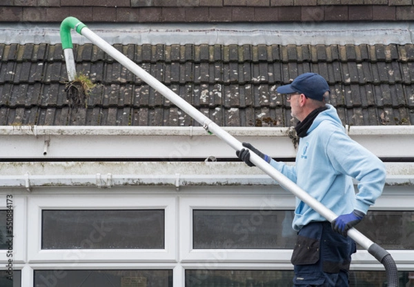 Fototapeta A window and gutter cleaner cleaning Dirty clogged white plastic pvc gutters and drain pipes with mossy green mould on plastic fascias. Blocked drains and guttering need  regular yard work maintenance