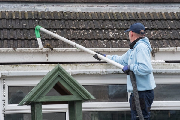 Fototapeta A window and gutter cleaner cleaning Dirty clogged white plastic pvc gutters and drain pipes with mossy green mould on plastic fascias. Blocked drains and guttering need  regular yard work maintenance