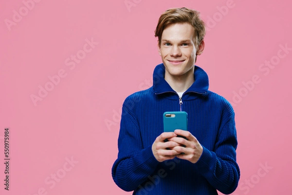Fototapeta a handsome red-haired guy stands with a phone in a blue case in his hands and smiles cutely at the camera