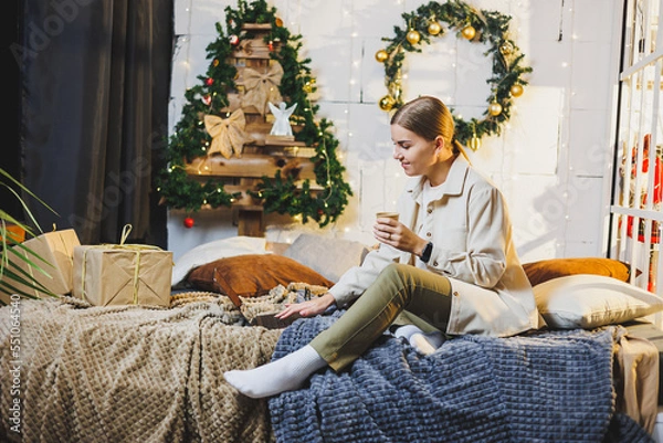 Fototapeta Young pretty woman with blond long hair in cozy clothes with coffee in hands using laptop watching movie and smiling on bed in room with Christmas tree at home. New Year holidays.