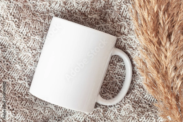 Fototapeta Mockup of white mug on knitted plaid with dried flowers, in neutral beige color. Blank coffee cup mug mock up in Boho style, flat lay.