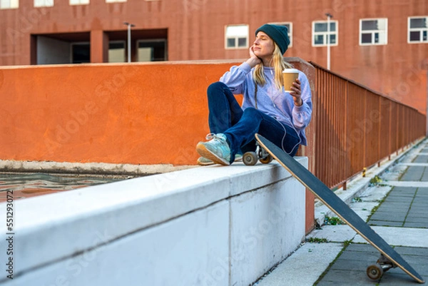 Fototapeta Young female teenager listens to music from her smartphone using headphones, relaxing moment of a skate girl, concept of street style and urban scene, Generation z people