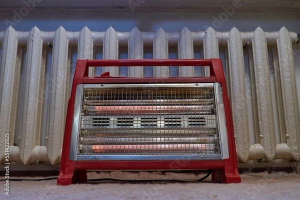 Fototapeta The red heater lies on the carpet in the bedroom. In the background is a cast iron radiator. The house has poor heating, you have to connect a working heating element, an additional source of heat.