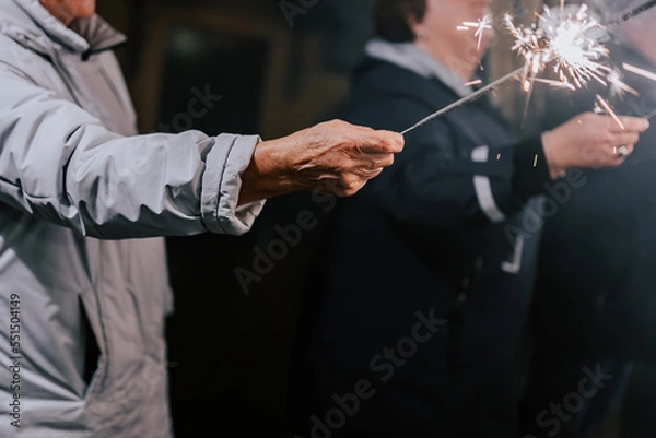 Fototapeta An old man holds a sparkling lights in his hand. Happy people with sparklers in their hands during celebration. Sparkler in hands on a wedding - guests holding lights in hand. 2023