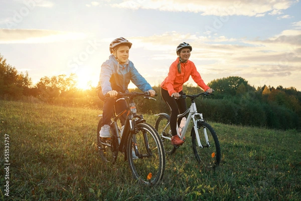 Fototapeta Mother and son ride a bike. Happy cute boy in helmet learn to riding a bike in park on green meadow in autumn day at sunset time. Family weekend. 4K video, Slow motion