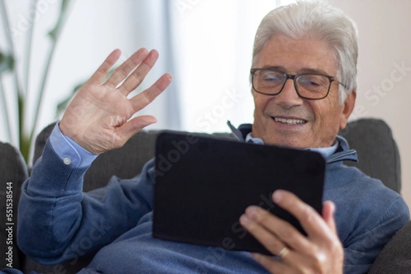 Fototapeta Smiling elderly man videoconferencing with family using digital tablet and waving hand. Positive gray-haired man sitting on sofa at home. Online chatting concept