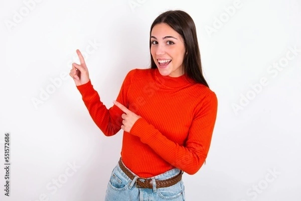 Fototapeta Young caucasian woman wearing red sweater over white background points at copy space indicates for advertising gives right direction