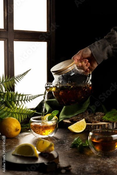Fototapeta Custard tea is poured from a glass teapot into a small cup. Hands in the frame. Black tea with lemon and mint