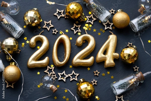 Fototapeta The golden figures 2024 made of candles on a black stone slate background are decorated with a festive decor of stars, sequins, fir branches, balls and garlands. Greeting card, happy New Year.