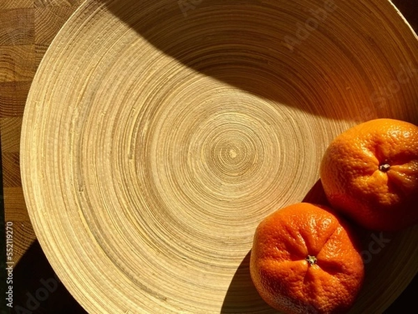 Fototapeta close-up of orange tangerines on a wooden kitchen plate in contrasting sunlight. orange tangerines on white wooden plate with a texture of concentric circles.