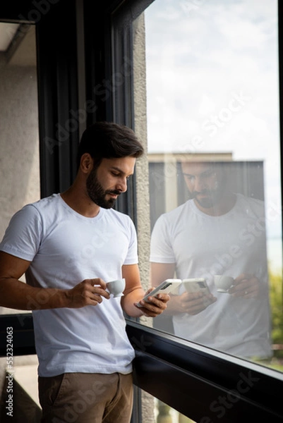 Obraz person using a phone, handsome boy looking at his phone next to a window in his room worried