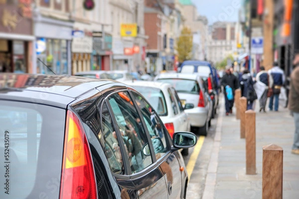Fototapeta A typical busy high street scene in the UK with parked cars and pedestrians.