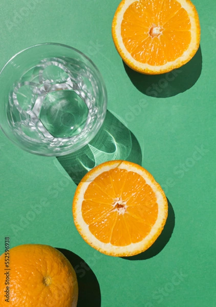 Fototapeta Composition with sliced orange fruit and empty glass on green background. Tropical summer minimal concept sunlit with sharp shadows