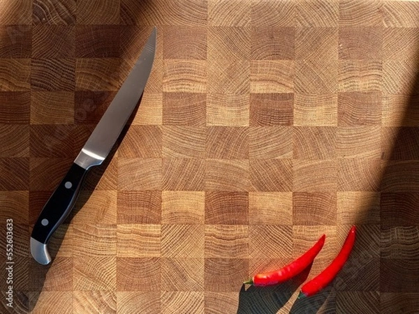 Fototapeta close-up of stainless kitchen knives and red hot chili peppers on the surface of a wooden board for cooking. Knives and red peppers on oak cutting board in bright rays of contrasting sunlight.