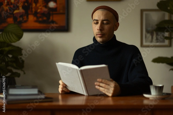 Fototapeta A caucasian man wearing a beanie and a knitted sweater sitting at a wooden table in a living room reading a book. 
