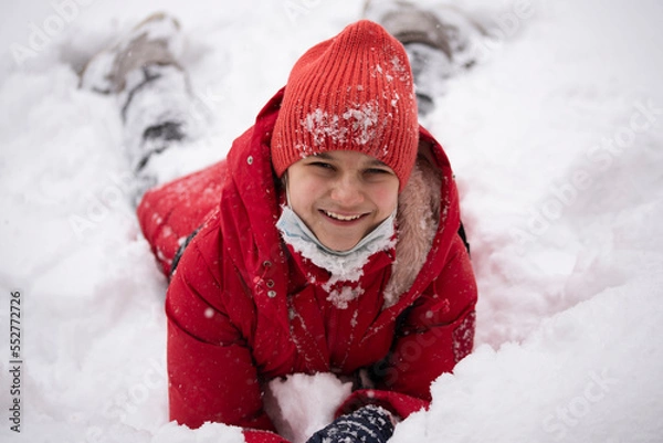 Fototapeta A girl in a red jacket in the snow. The girl smiles and lies on the snow. Winter fun.