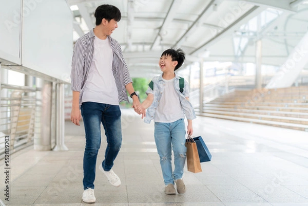 Obraz Young Asia boy holding colorful shopping bag and walking with father in the shopping central area. Enjoyment on shopping and happy with gift during end of the year. Mid year sale and holiday concept