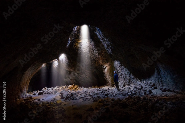 Fototapeta Adventurous athletic male standing in a lava tube looking at the sunlight shinning down into the cave through holes in the ceiling.

