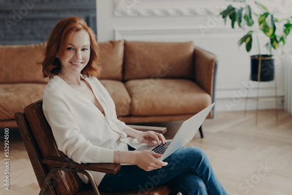 Fototapeta Sideways shot of woman entrepreneur sits in comfortable armchair in modern apartment with luxury furtniture, communicates with colleague through online video chat, uses laptop free internet connection