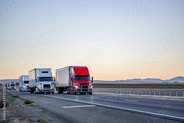 Obraz Convoy of industrial big rigs semi trucks with semi trailers driving with cargo on the straight highway road at twilight time